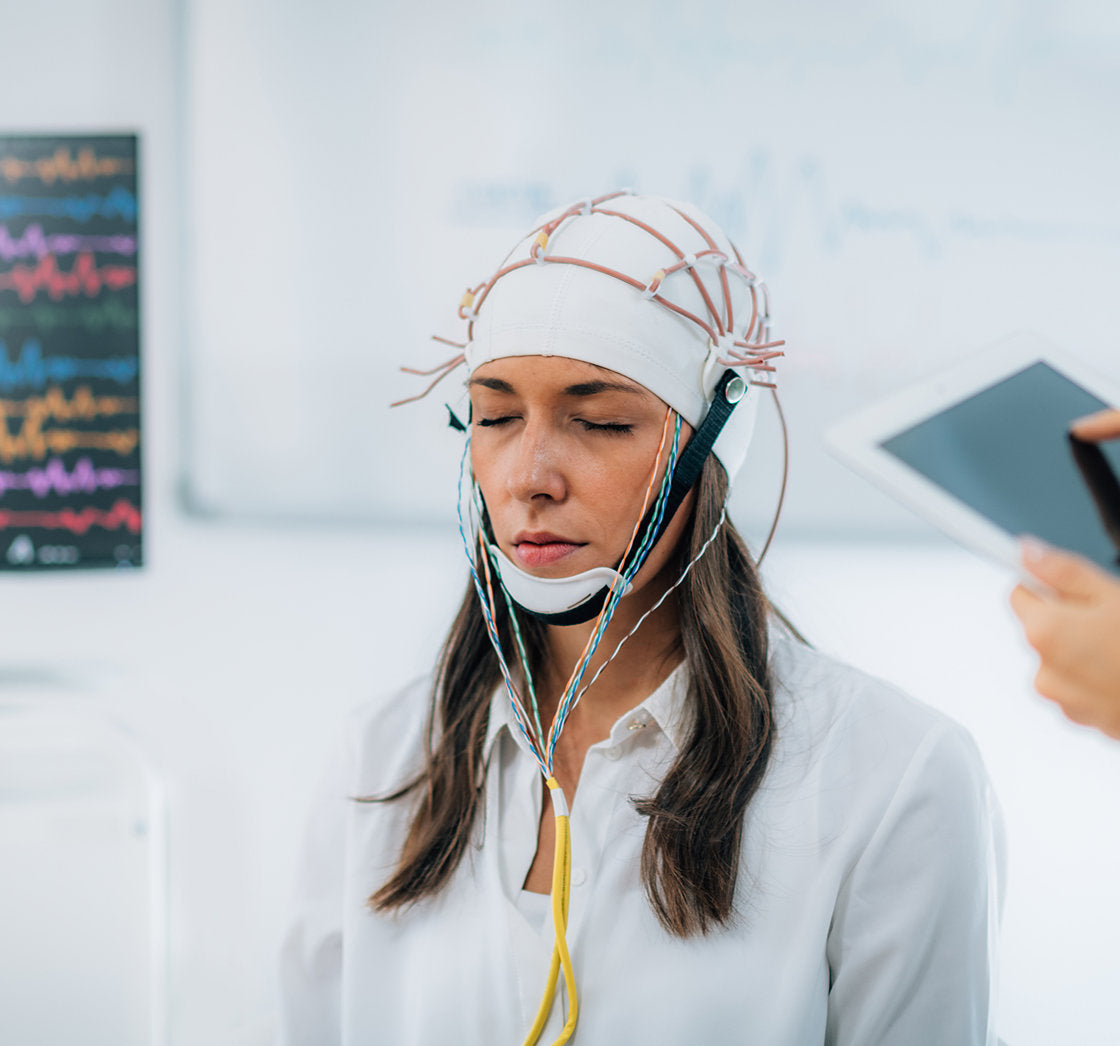 A person wearing an EEG cap with electrodes is being monitored for brain activity in a clinical setting. A healthcare professional holds a tablet, while a monitor in the background displays colorful brain wave patterns.